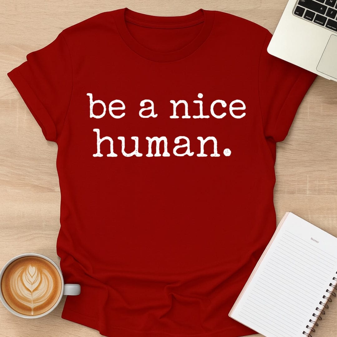 Red t-shirt with 'be a nice human' text on a wooden surface with a laptop, coffee, and notebook.