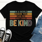 Black Slogan t-shirt with motivational quote 'In a world where you can be anything, be kind' on a white background with sneakers and jeans.